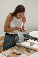 Taking picture of handmade ceramics on workshop. Pottery blog owner making photo of clay plate on smartphone working in studio. Woman blogger artist sharing process of crockery creation