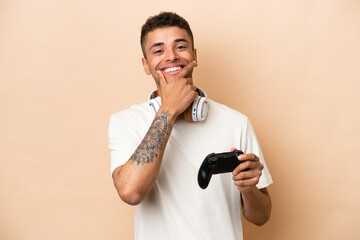 Young Brazilian man playing with a video game controller isolated on beige background happy and smiling