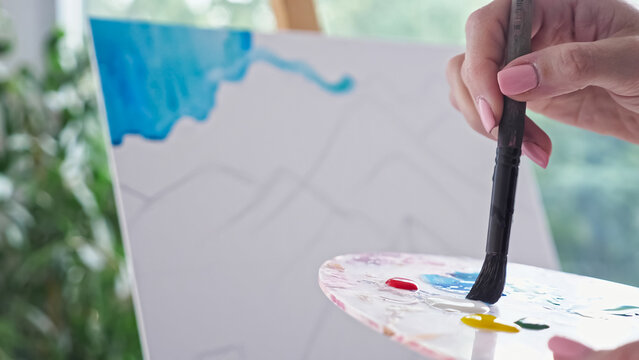 Professional artist paints mountain landscape using paintbrush. Person hand soaks paintbrush into colour on palette and paints sky on canvas closeup