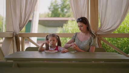 Mother opens pack with sauce dining with little daughter on terrace of restaurant slow motion. Woman with girl rests in street cafe. Family lunch