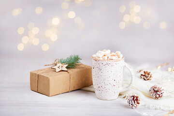 Christmas holiday background, greeting card. A mug of cocoa or hot chocolate with marshmallows, a gift in kraft paper on the background of a beautiful bokeh. 