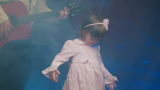 Little girl on stage in pink vintage dress, she dances, her father plays acoustic guitar. Color music is shining and smoke is billowing. Father Day. Performance on stage. Hobbies and creativity