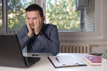 Young man disappointed while working on laptop. He is sitting at table at home. Worried remote...