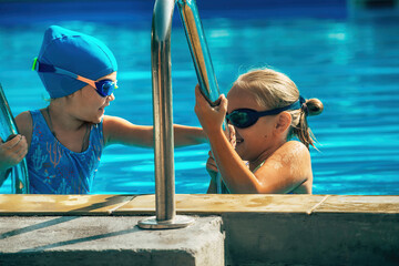 Two little cheerful girls sportswomen in waterproof goggles and swimsuits at a swimming class in an outdoor pool on a warm sunny day