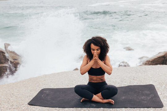 Young woman sitting on the beach meditating with eyes closed. Female with hands together and waves behind doing yoga.