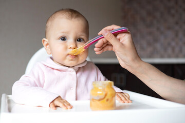 Baby food. A baby in a high chair eats vegetable puree from a spoon. Mom feeds the baby from a jar. Kitchen. Lifestyle.