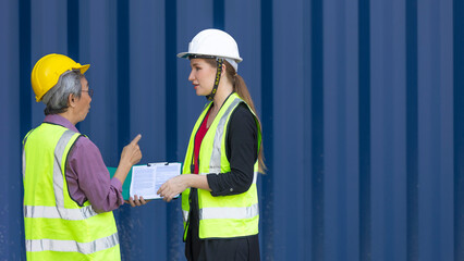 Elderly male engineer with safety vest and helmet provides advice and support to young female worker next to blue shipping container. Elder manager is training young staff in a logistics place