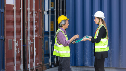 Elderly male engineer with safety vest and helmet provides advice and support to young female worker next to blue shipping container. Elder manager is training young staff in a logistics place