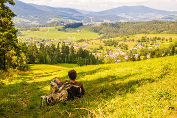 Młody mężczyzna z plecakiem siedzący na skraju lasu patrzący na piękny krajobraz w górach - obrazy, fototapety, plakaty