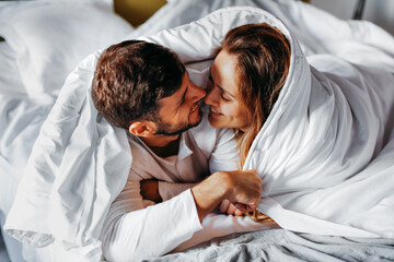 Cheerful young couple caressing under white blanket in bed