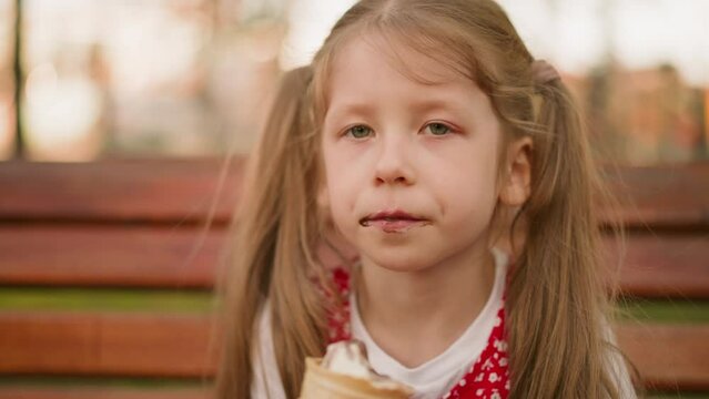 Preschooler girl with ponytails eats ice cream with pleasure sitting on wooden bench closeup. Kid bites cold dessert at sunset. Delicious sundae snack