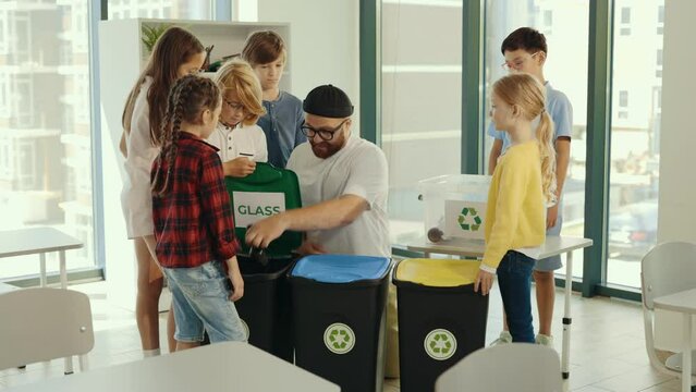 Male teacher shows to the pupils how and where sort rubbish correctly and sorting the rubbish on the garbage sorting class together with pupils. Ecological lesson. Bsck to school. Education.