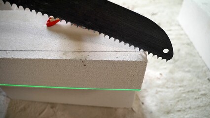 A construction worker saws an aerated concrete block with a hand saw at a construction site. Builder using a hacksaw cuts aerated concrete block. Construction contractor cuts autoclaved