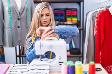 Blonde woman dressmaker designer using sew machine checking the time on wrist watch, relaxed and confident