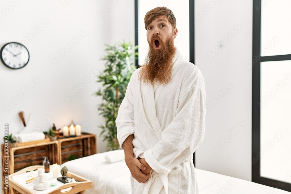 Wall mural redhead man with long beard wearing bathrobe at wellness spa scared and amazed with open mouth for s