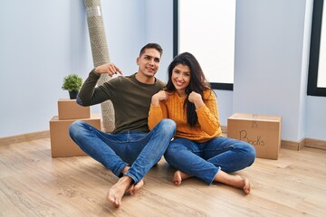 Young couple sitting on the floor at new home looking confident with smile on face, pointing oneself with fingers proud and happy.