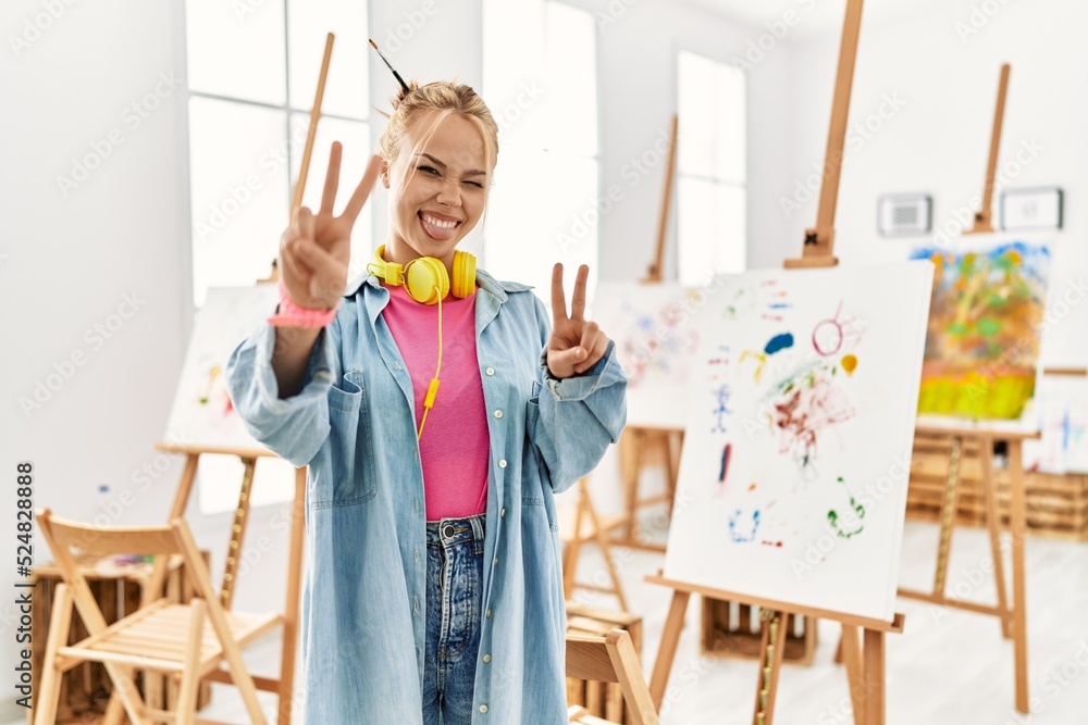 Poster Young caucasian girl at art studio smiling with tongue out showing fingers of both hands doing victory sign. number two.