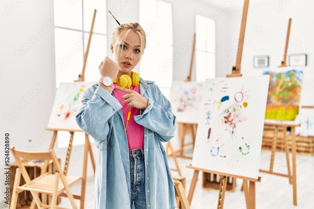 Poster Young caucasian girl at art studio in hurry pointing to watch time, impatience, looking at the camera with relaxed expression