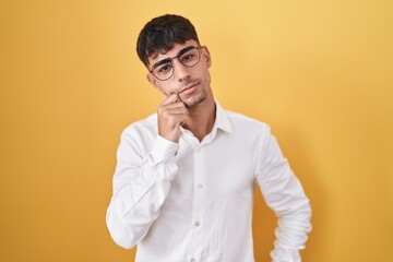 Fototapeta premium Young hispanic man standing over yellow background with hand on chin thinking about question, pensive expression. smiling with thoughtful face. doubt concept.