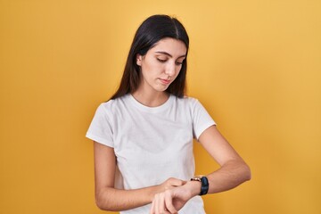 Young beautiful woman standing over yellow background checking the time on wrist watch, relaxed and confident