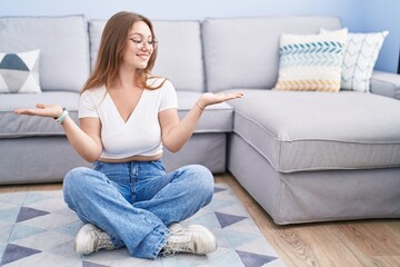 Young caucasian woman sitting on the floor at the living room smiling showing both hands open...