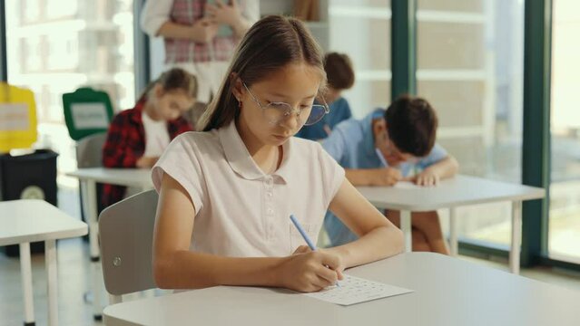Beautiful long-haired brunette asian pupil wearing glasses sits at a first desk in the classroom and fills out tests thinks about answers. Elementary school. Learning and education concept.