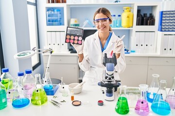 Fototapeta premium Young blonde woman working at scientist laboratory with make up smiling happy pointing with hand and finger to the side