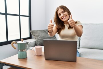 Fototapeta na wymiar Young brunette woman using laptop at home drinking a cup of coffee success sign doing positive gesture with hand, thumbs up smiling and happy. cheerful expression and winner gesture.