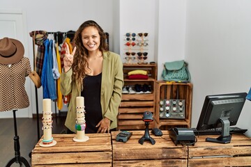 Beautiful hispanic woman working at fashion shop showing and pointing up with fingers number two while smiling confident and happy.