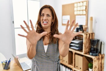 Middle age hispanic woman at the office afraid and terrified with fear expression stop gesture with...
