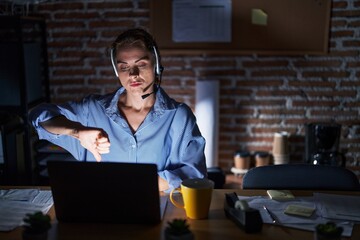 Beautiful brunette woman working at the office at night looking unhappy and angry showing rejection and negative with thumbs down gesture. bad expression.