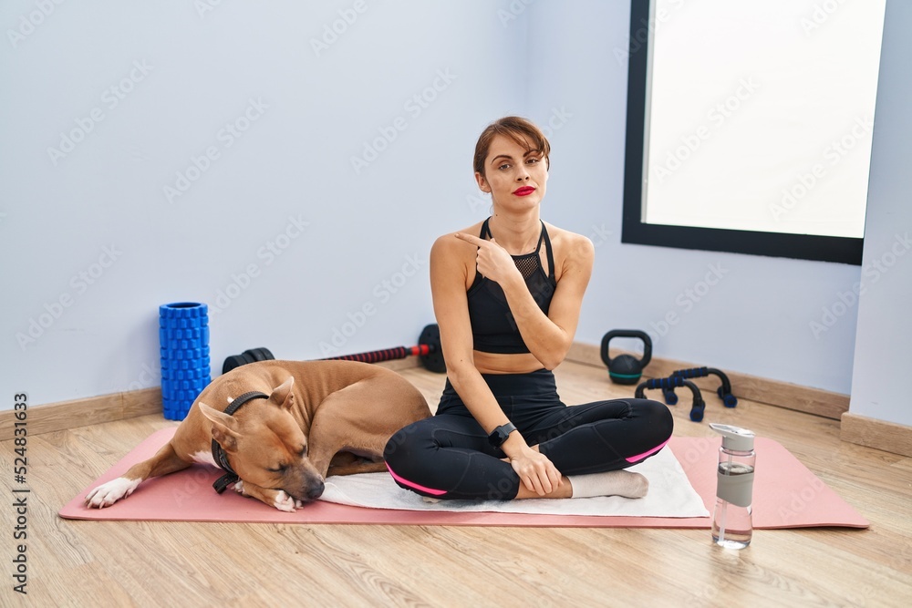 Sticker Young beautiful woman sitting on yoga mat pointing with hand finger to the side showing advertisement, serious and calm face