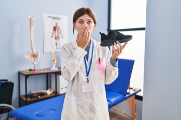 Young physiotherapist woman holding sneakers covering mouth with hand, shocked and afraid for mistake. surprised expression