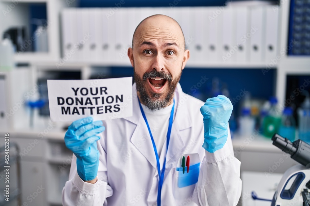 Wall mural Young hispanic man working at scientist laboratory holding you donation matters banner screaming proud, celebrating victory and success very excited with raised arms