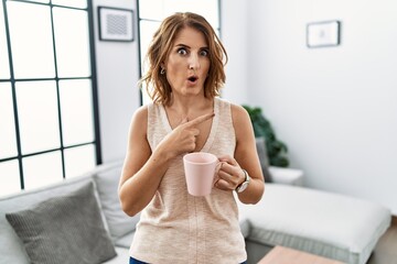 Middle age woman drinking a cup coffee at home surprised pointing with finger to the side, open mouth amazed expression.