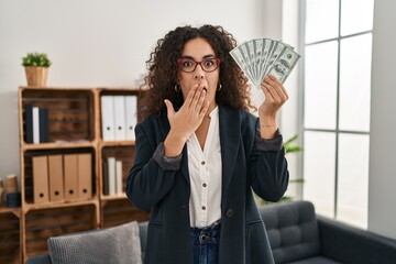 Young hispanic woman holding dollars covering mouth with hand, shocked and afraid for mistake....