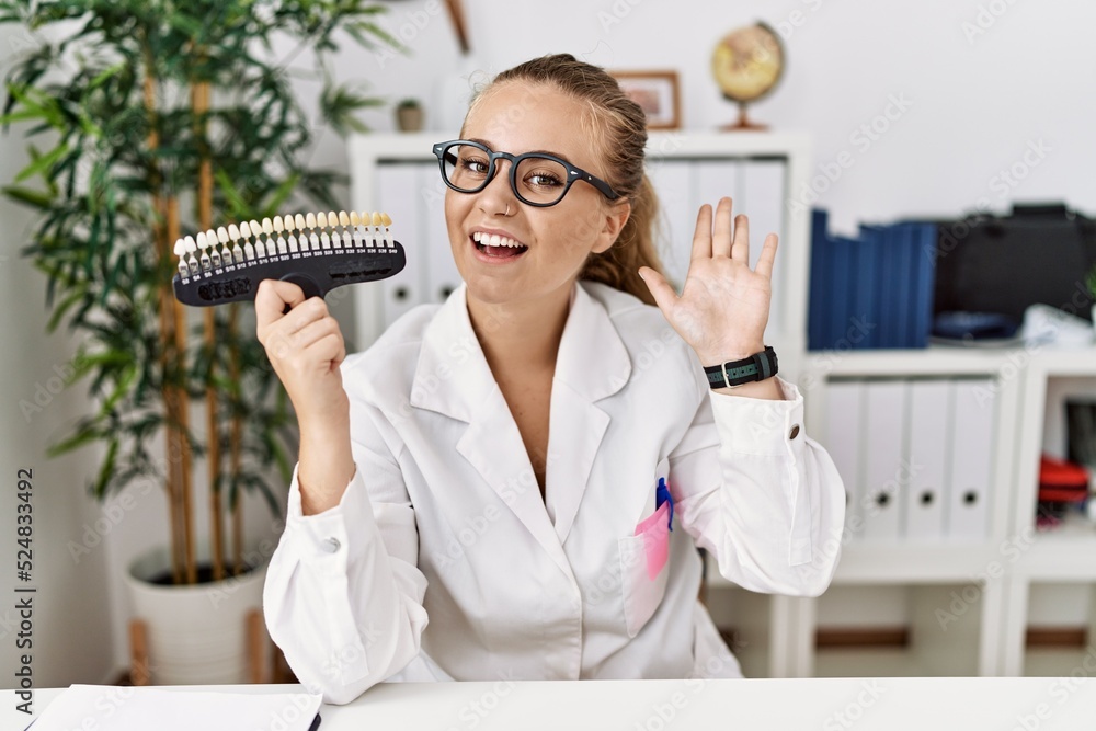 Poster Young caucasian woman holding teeth whitening palette waiving saying hello happy and smiling, friendly welcome gesture