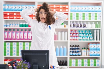 Hispanic woman with curly hair working at pharmacy drugstore crazy and scared with hands on head,...