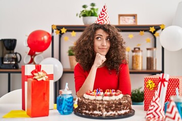 Hispanic woman with curly hair celebrating birthday holding big chocolate cake serious face thinking about question with hand on chin, thoughtful about confusing idea