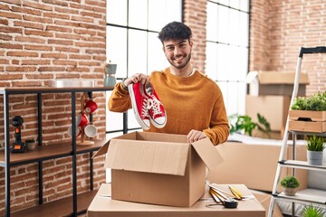 Young hispanic man smiling confident holding sneakers unpacking cardboard box at new home
