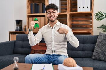 Hispanic man with beard holding support green ribbon at therapy clinic pointing finger to one self smiling happy and proud