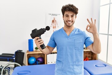 Naklejka na ściany i meble Hispanic physiotherapy man working at pain recovery clinic with muscle gun doing ok sign with fingers, smiling friendly gesturing excellent symbol