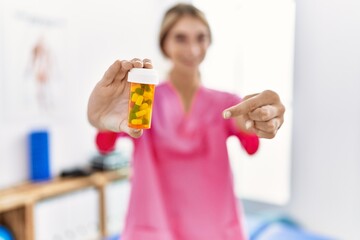 Young physiotherapist woman working at pain recovery clinic holding pills smiling happy pointing with hand and finger