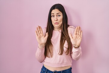 Young brunette woman standing over pink background moving away hands palms showing refusal and...