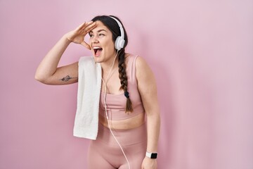 Young brunette woman wearing sportswear and headphones very happy and smiling looking far away with hand over head. searching concept.