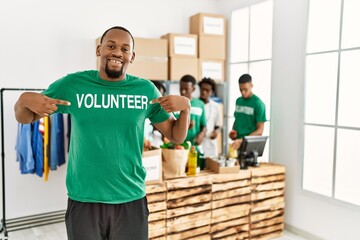 Group of young african american volunteers working at charity center. Man smiling happy and pointing with fingers to volunteer uniform.
