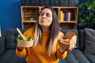 Young hispanic woman holding healthy salad and fried chicken wings looking at the camera blowing a...