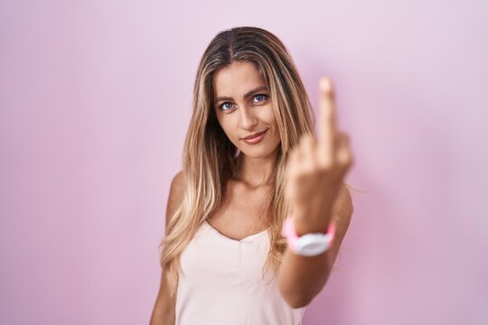 Young blonde woman standing over pink background showing middle finger, impolite and rude fuck off expression