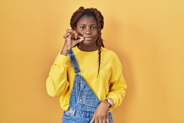 African woman standing over yellow background mouth and lips shut as zip with fingers. secret and...