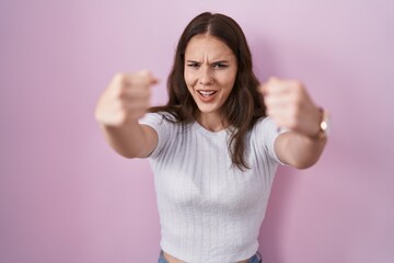 Young hispanic girl standing over pink background angry and mad raising fists frustrated and furious while shouting with anger. rage and aggressive concept.